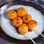 Two homemade tofu mitarashi dango on an off-white plate with brown rim