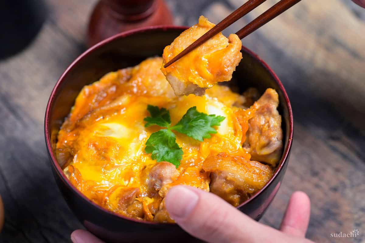 Yuto Omura holding a bowl of oyakodon (chicken and egg rice bowl) in one hand, and chopsticks holding up a piece of chicken over the bowl