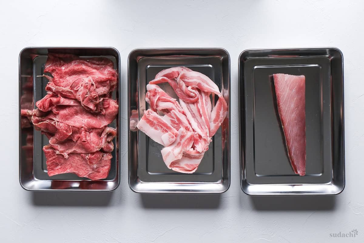 Thinly sliced wagyu beef, pork belly and a yellowtail block in steel rectangular containers side by side (for shabu shabu protein options)