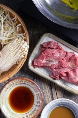 Ingredients for shabu shabu hot pot laid out on a table including ponzu dipping sauce, sesame dipping sauce, vegetables in a bamboo woven basket, thinly sliced wagyu beef on a plate and kombu dashi in a stainless steel pot on a tabletop stove