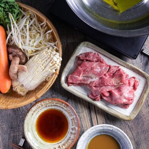 Ingredients for shabu shabu hot pot laid out on a table including ponzu dipping sauce, sesame dipping sauce, vegetables in a bamboo woven basket, thinly sliced wagyu beef on a plate and kombu dashi in a stainless steel pot on a tabletop stove