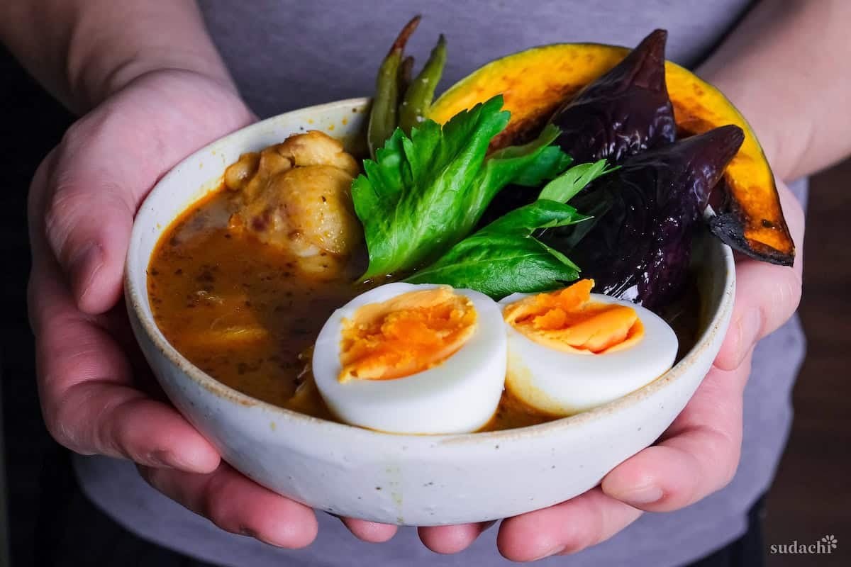 Yuto Omura holding Japanese Soup Curry topped with oven roasted vegetables, boiled eggs and a celery leaf in a cream bowl