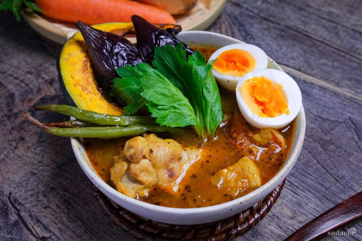 Japanese Soup Curry topped with oven roasted vegetables, boiled eggs and a celery leaf in a cream bowl