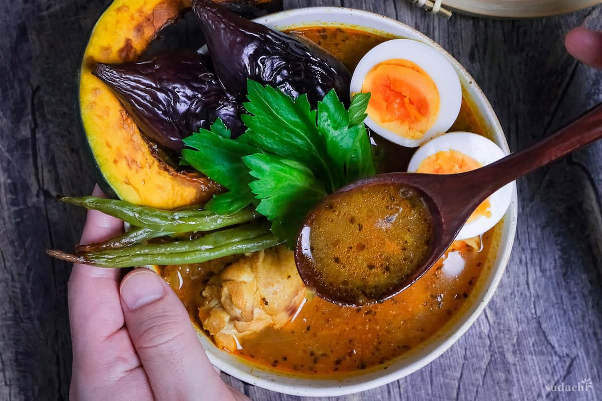 Japanese Soup Curry topped with oven roasted vegetables, boiled eggs and a celery leaf in a cream bowl with a dark wooden spoon held above it