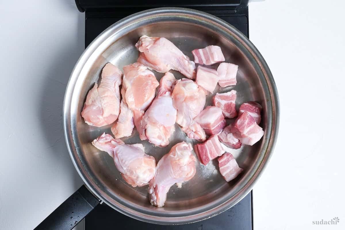 Searing chicken drumsticks and cubes of pork bell in a stainless steel pan on the stove top