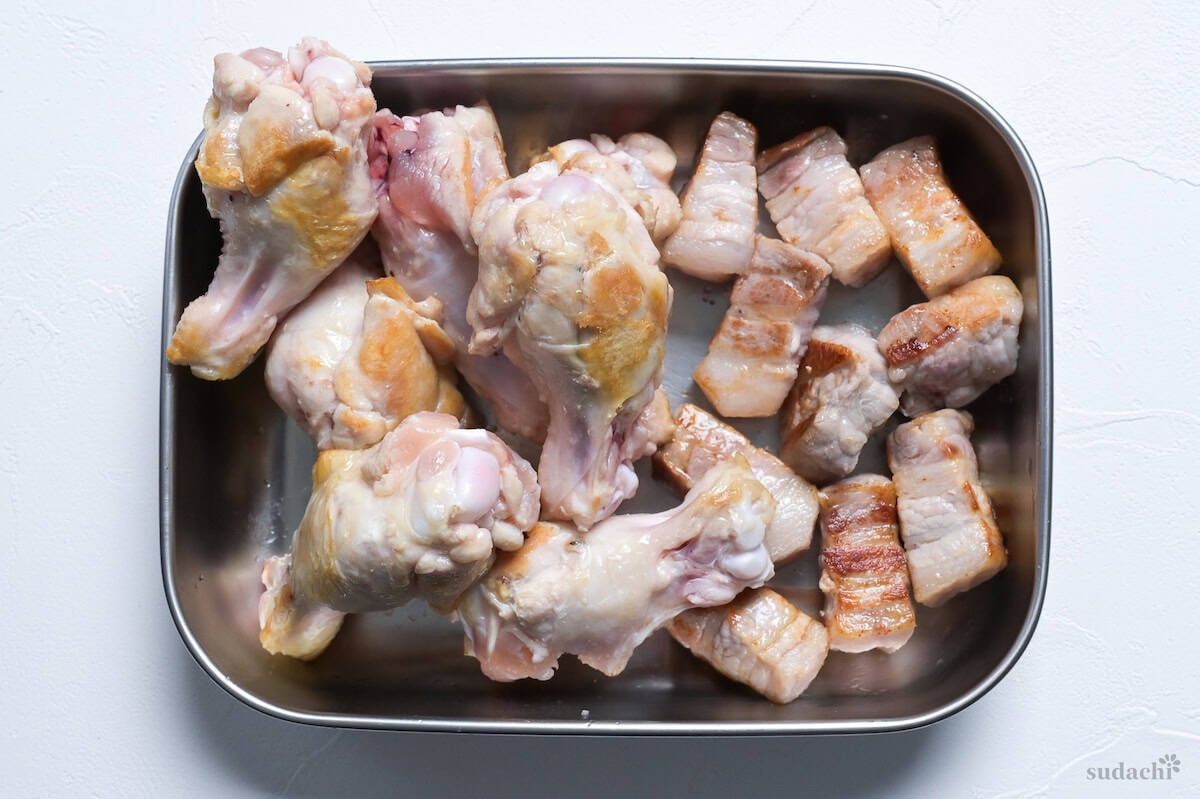 Seared chicken drumsticks and pork belly cubes in a steel container