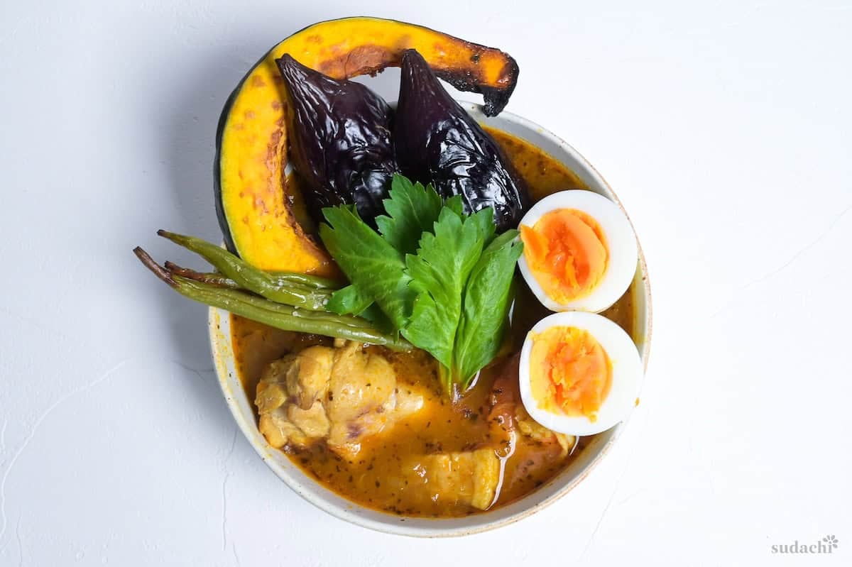 Japanese Soup Curry topped with oven roasted vegetables, boiled eggs and a celery leaf in a cream bowl
