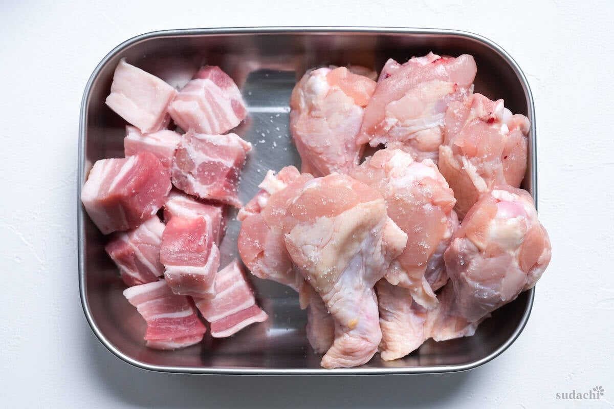 Cubes of pork belly and chicken drumsticks sprinkled with salt in a steel container