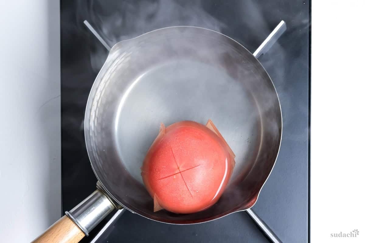 One whole tomato with a cross cut on top, in a pan of hot water on the stove top