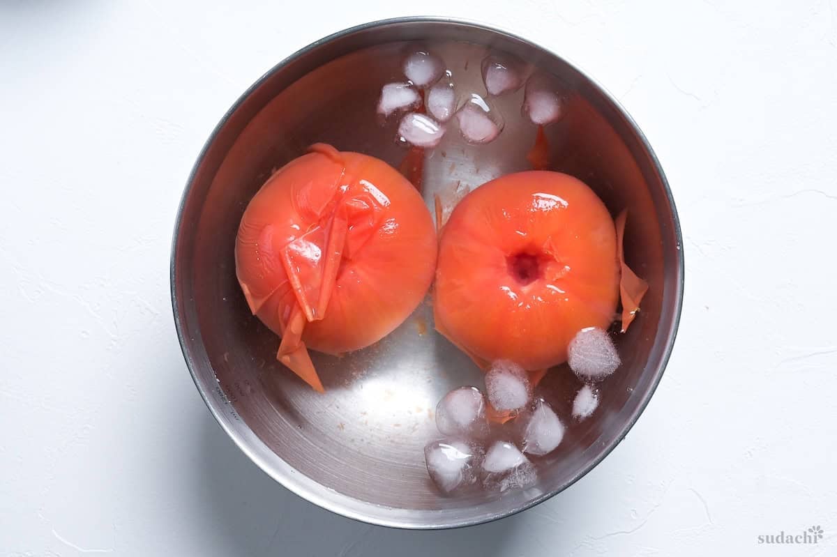 two blanched tomatoes in a bowl of ice water with skin peeling off
