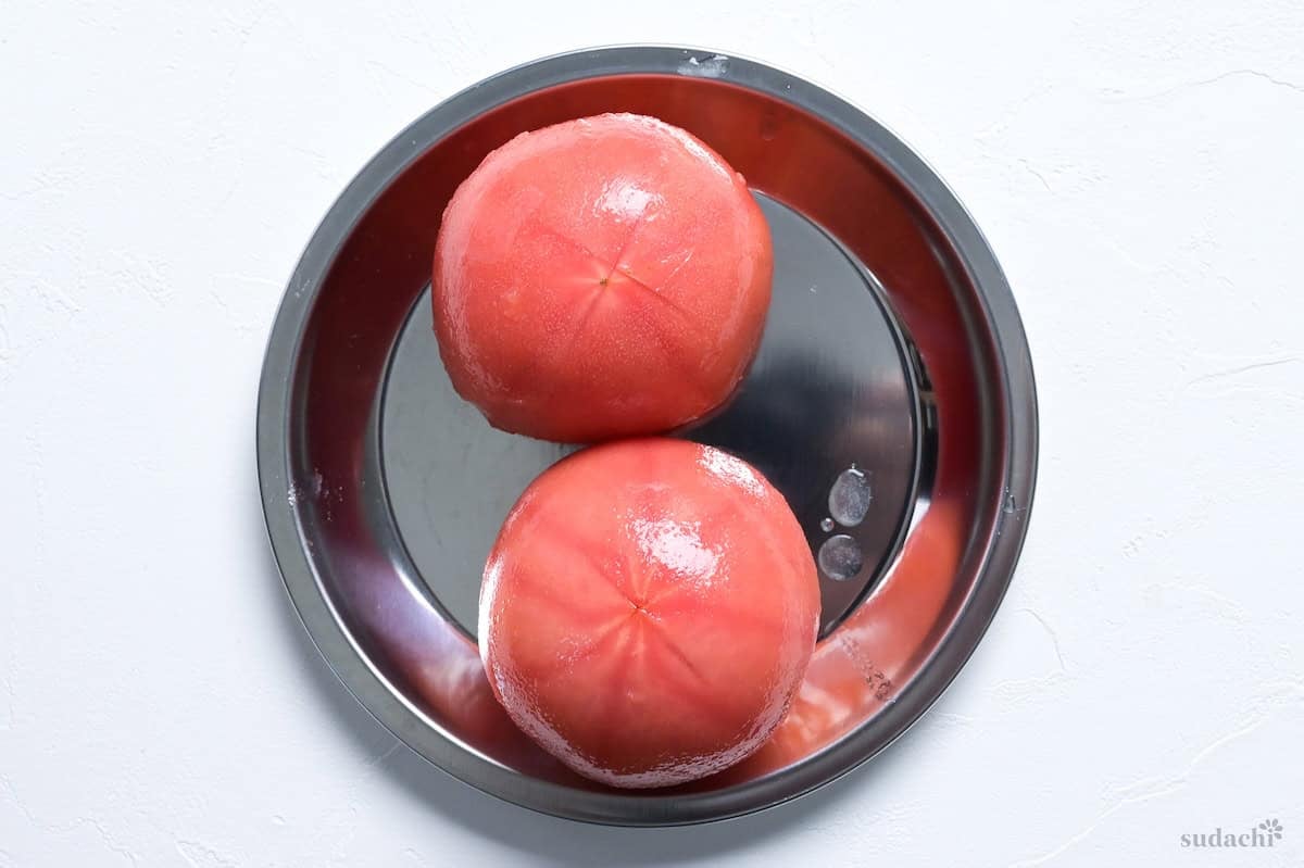 Two blanched and peeled whole tomatoes on a steel prep plate on a white background