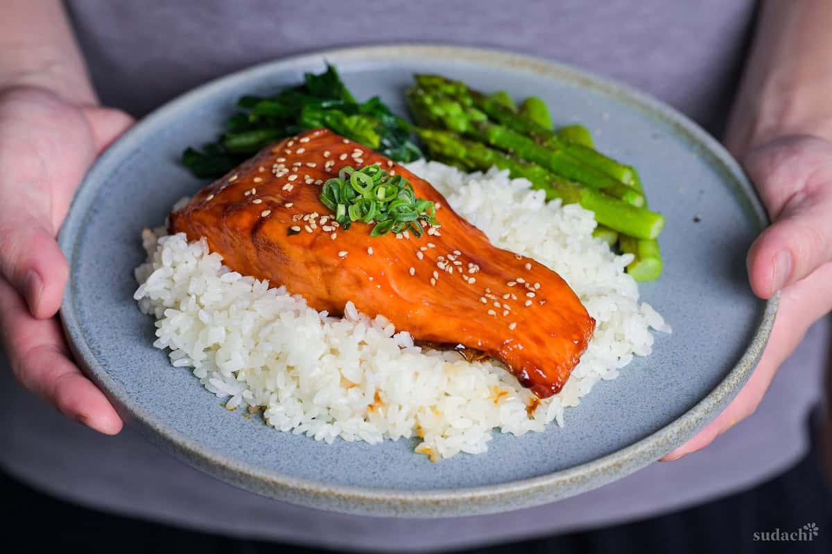 Yuto Omura holding up Teriyaki Salmon sprinkled with toasted sesame seeds and chopped green onions served over white rice on a blue plate with sautéed asparagus and wilted spinach