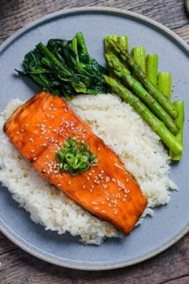 Teriyaki Salmon sprinkled with toasted sesame seeds and chopped green onions served over white rice on a blue plate with sautéed asparagus and wilted spinach top down