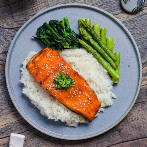 Teriyaki Salmon sprinkled with toasted sesame seeds and chopped green onions served over white rice on a blue plate with sautéed asparagus and wilted spinach top down
