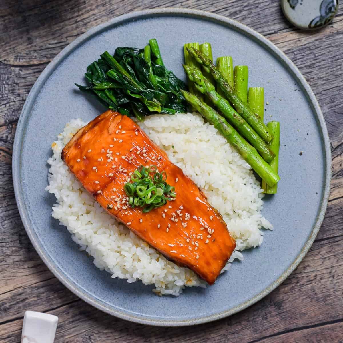 Teriyaki Salmon sprinkled with toasted sesame seeds and chopped green onions served over white rice on a blue plate with sautéed asparagus and wilted spinach top down