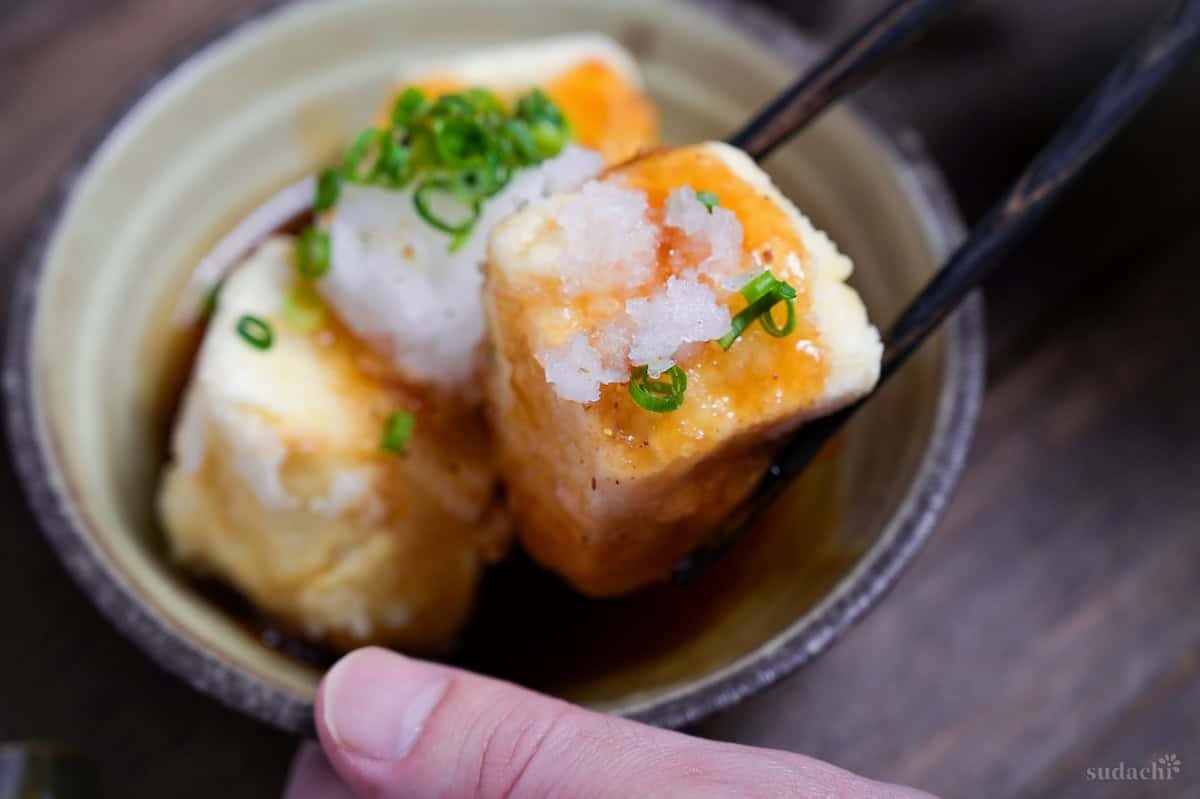 Agedashi tofu topped with grated daikon radish and chopped green onion, with black chopsticks holding up one piece