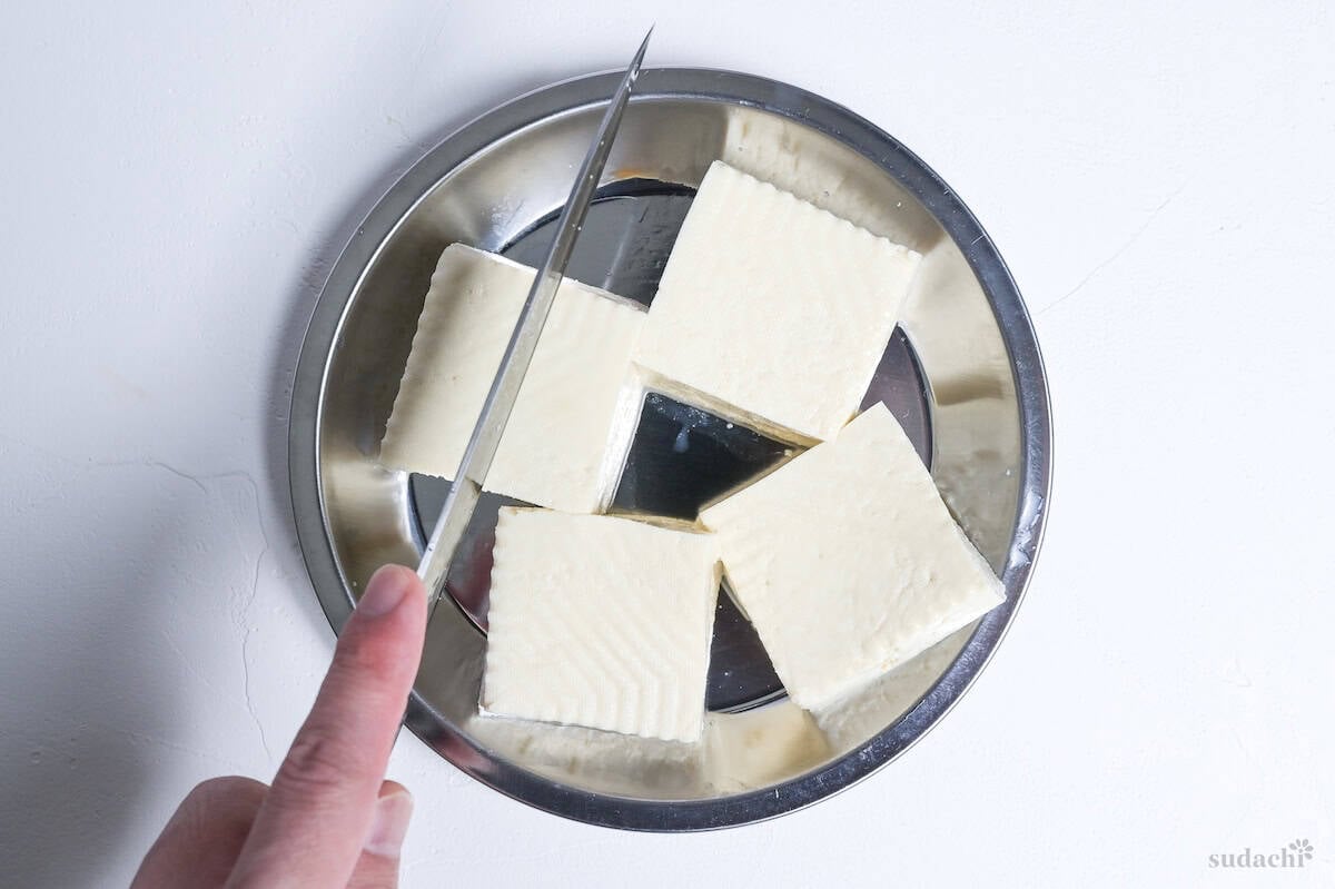 Four pieces of pressed firm tofu on a silver plate with a hand holding a knife above them