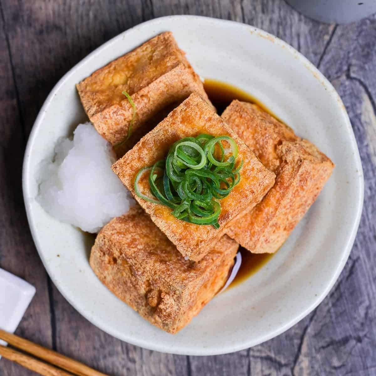 Four pieces of atsuage tofu in a cream bowl with dashi soy sauce and grated daikon radish