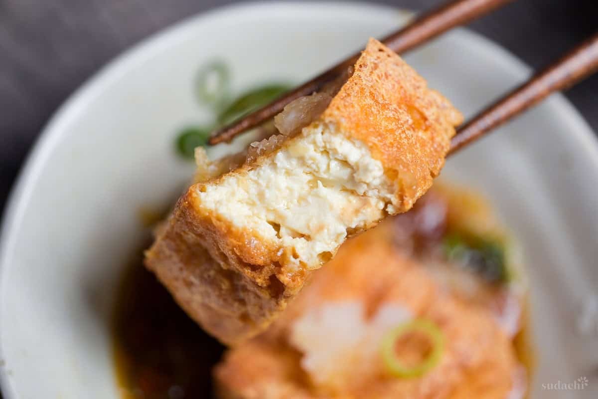 A piece of atsuage tofu with a bite missing to reveal inside, held up with wooden chopsticks