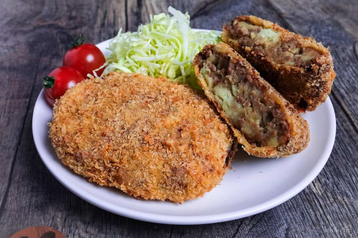 Two Japanese meat and potato korokke (croquettes) with one cut in half showing inside, on a white plate next to shredded cabbage and two mini tomatoes (side view)