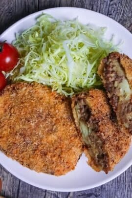 Two Japanese meat and potato korokke (croquettes) with one cut in half showing inside, on a white plate next to shredded cabbage and two mini tomatoes top down thumbnail