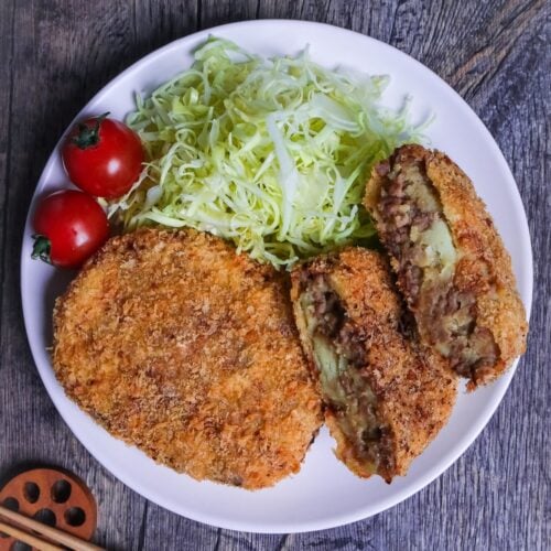 Two Japanese meat and potato korokke (croquettes) with one cut in half showing inside, on a white plate next to shredded cabbage and two mini tomatoes top down thumbnail