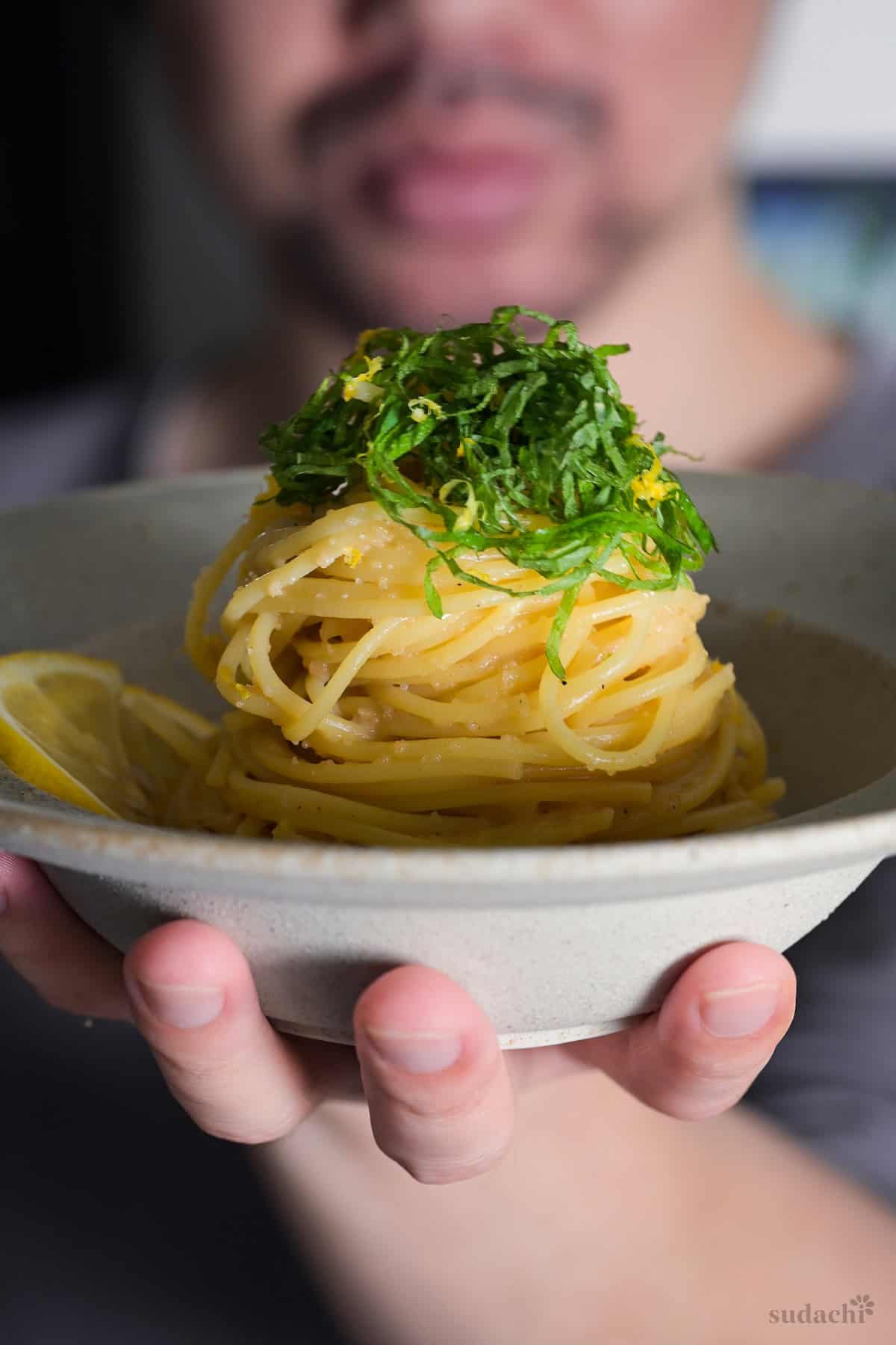 Yuto Omura holding up mentaiko pasta topped with shredded shiso leaves and lemon zest in a cream-colored ceramic pasta dish