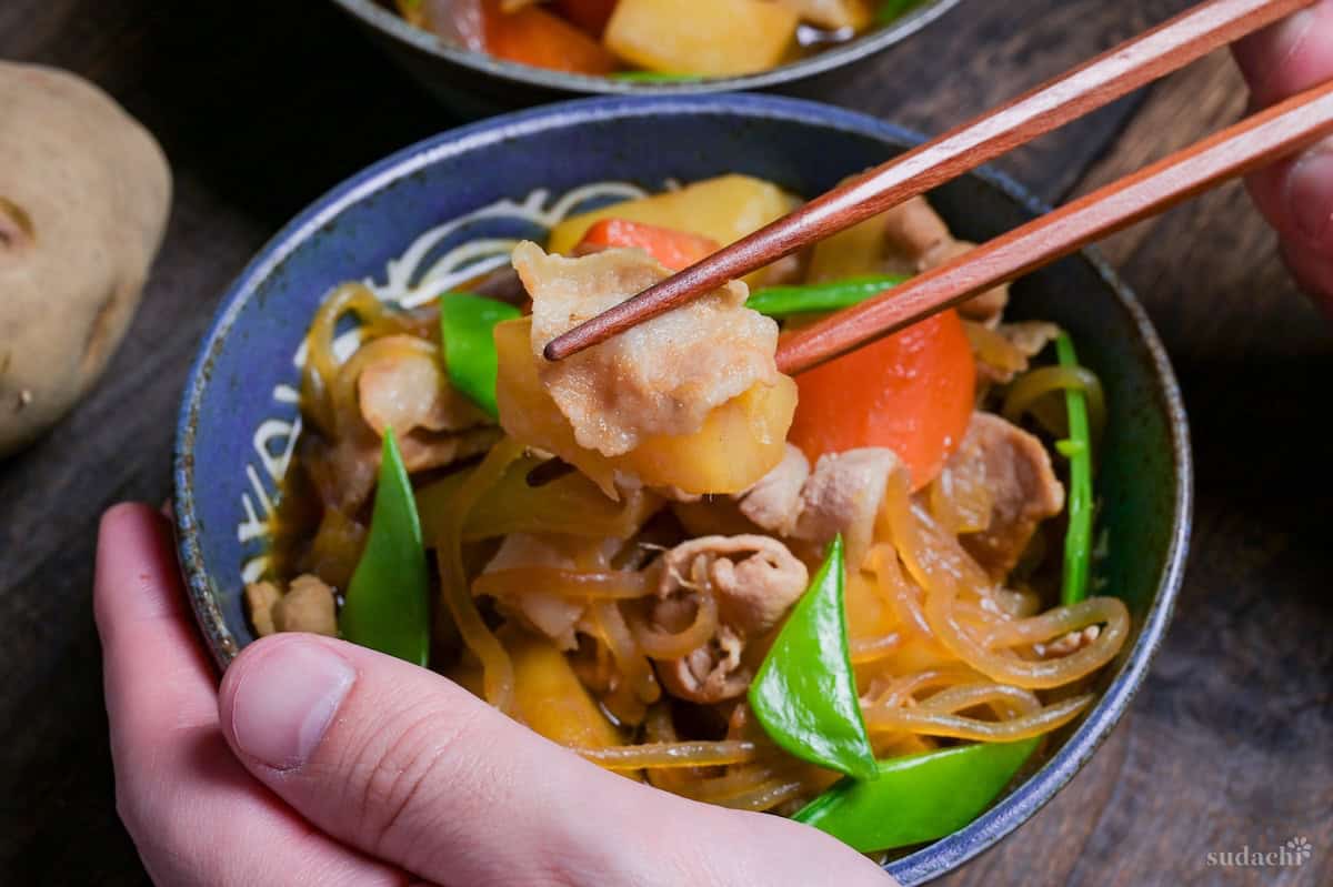 Nikujaga (Japanese meat and potato stew) with blanched snow peas in a blue bowl, with wooden chopsticks holding up a piece of pork and potato