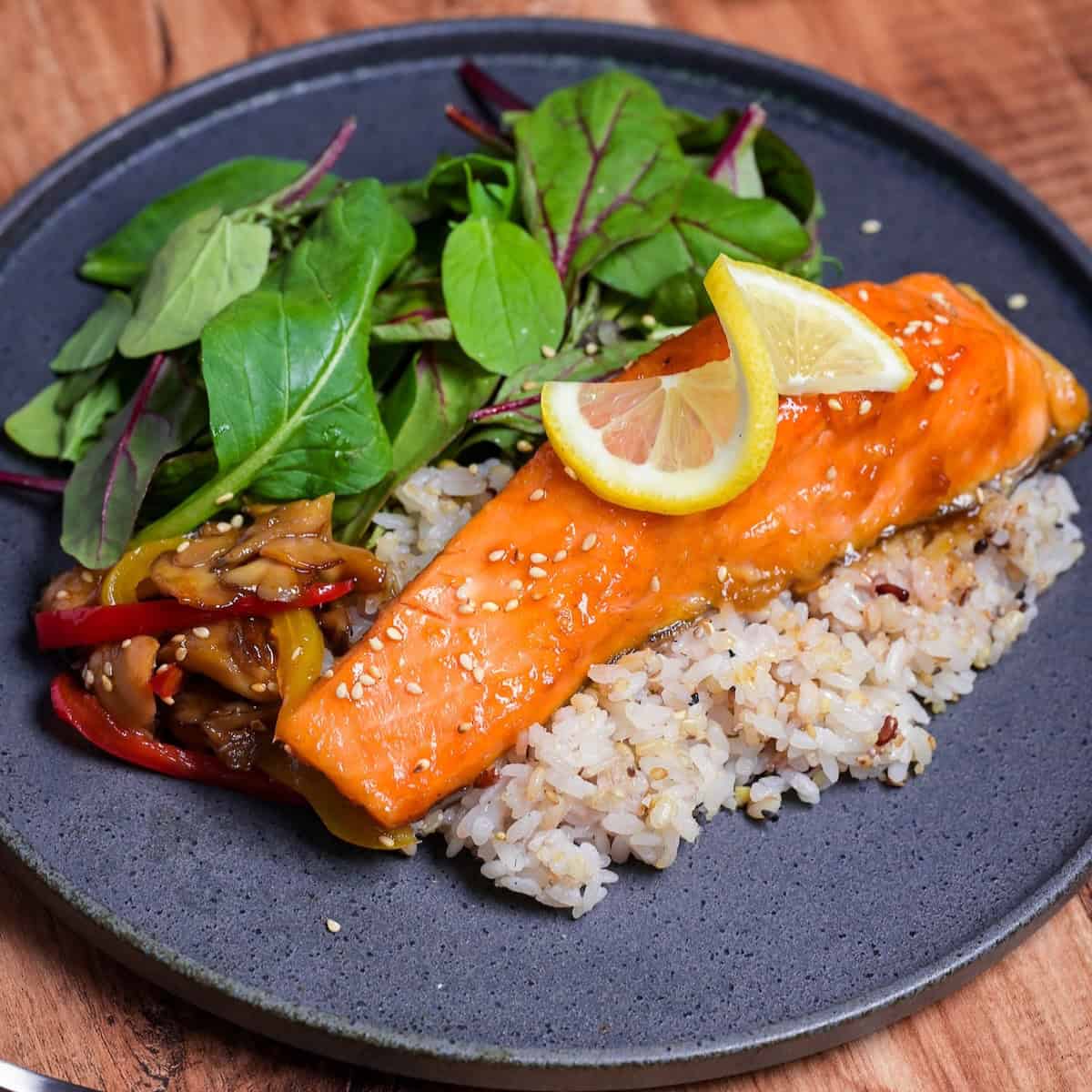 ponzu salmon on a bed of multigrain rice next to sautéed vegetables and baby leaf salad on a gray plate