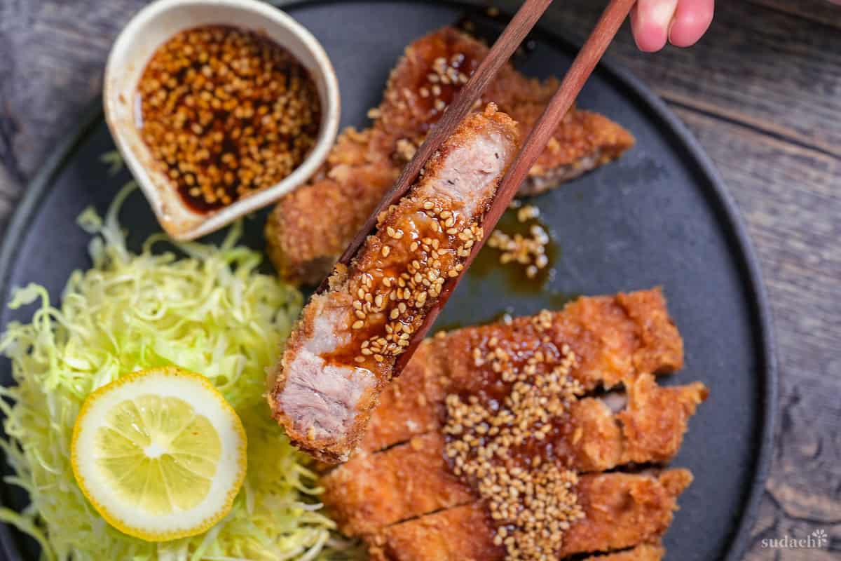 wooden chopsticks holding up a piece of tonkatsu (Japanese pork cutlet) drizzled with homemade tonkatsu sauce