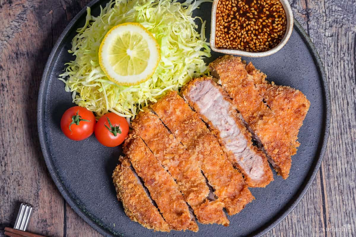 Homemade Tonkatsu Pork Cutlet cut into slices and served next to shredded cabbage, mini tomatoes and a small jug of homemade tonkatsu sauce on a dark gray plate
