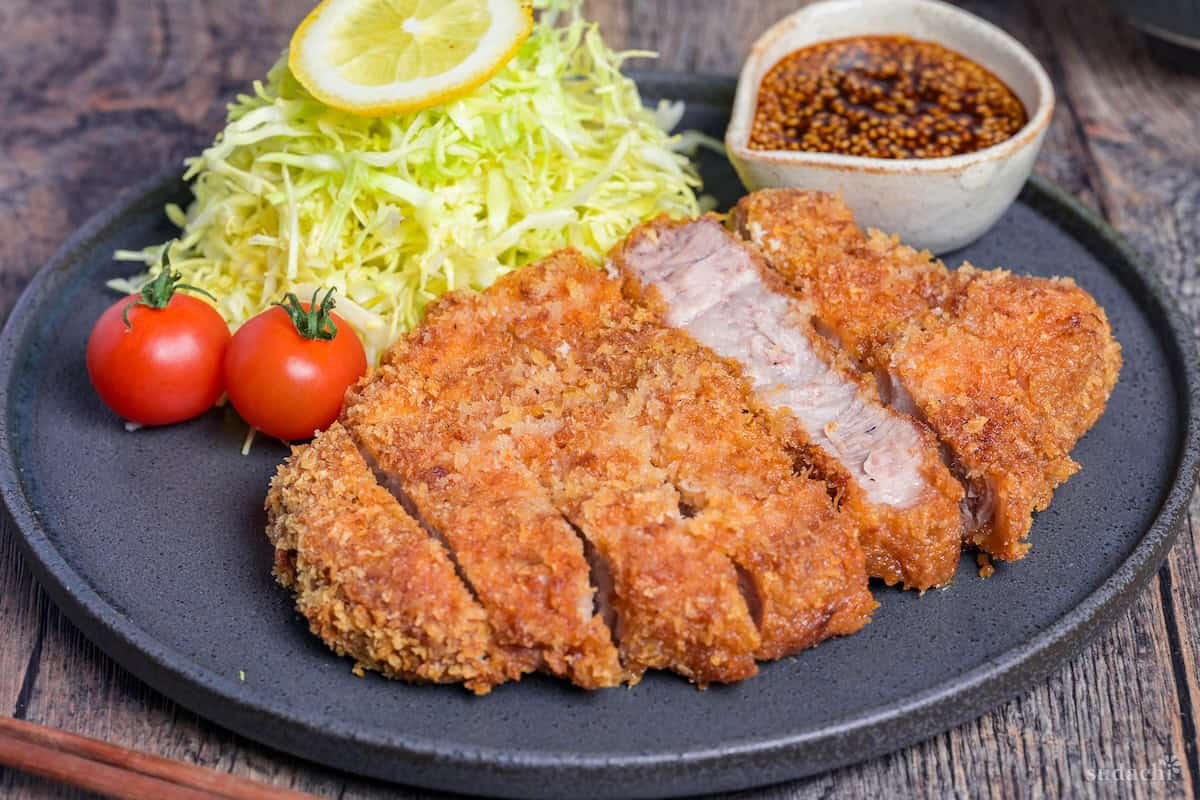 Homemade Tonkatsu Pork Cutlet cut into slices and served next to shredded cabbage, mini tomatoes and a small jug of homemade tonkatsu sauce on a dark gray plate side view