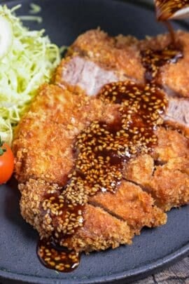Homemade Tonkatsu Pork Cutlet cut into slices, drizzled with homemade sauce and served next to shredded cabbage and mini tomatoes on a dark gray plate
