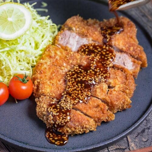 Homemade Tonkatsu Pork Cutlet cut into slices, drizzled with homemade sauce and served next to shredded cabbage and mini tomatoes on a dark gray plate