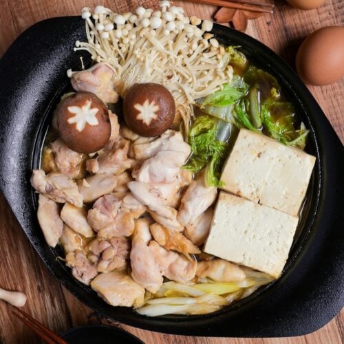 Chicken sukiyaki with enoki mushrooms, shiitake, cabbage and tofu in a cast iron pot, top down view