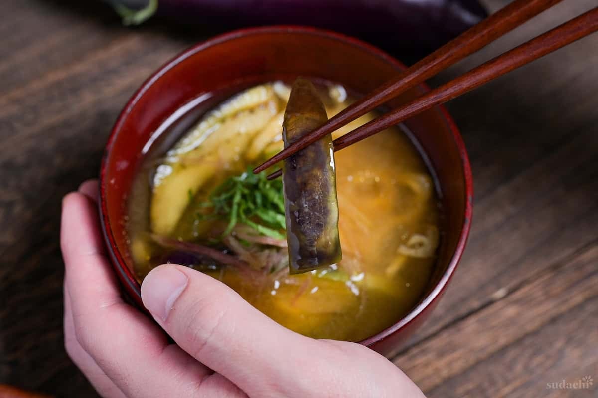 eggplant miso soup topped with Japanese ginger and shredded Perilla leaves in a red bowl with wooden chopsticks holding up a piece of eggplant