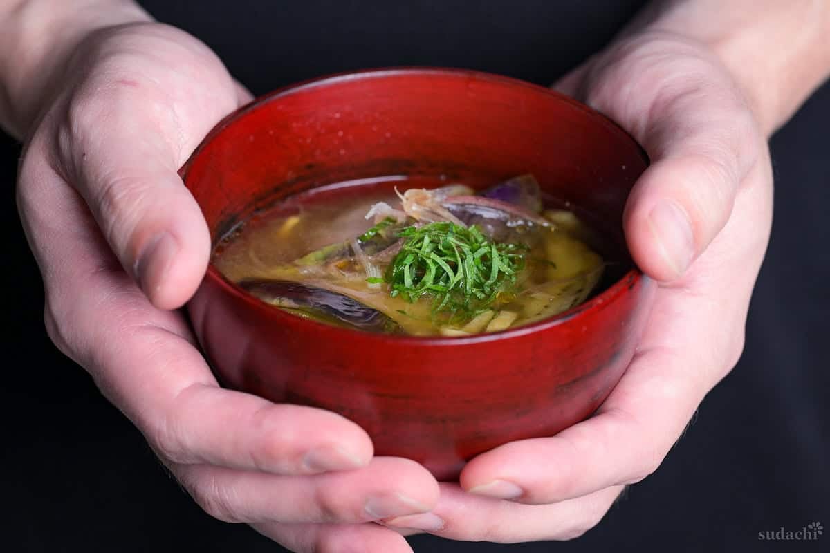 Yuto Omura holding up eggplant miso soup topped with Japanese ginger and shredded Perilla leaves in a red bowl