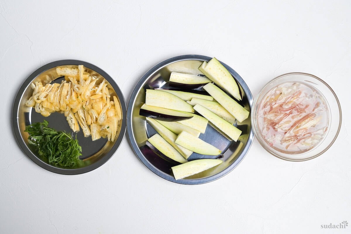 Prep for eggplant miso soup: shredded tofu pouches and perilla leaves on a silver plate, eggplant cut into sticks on another plate, and shredded myoga (Japanese ginger) soaking in a glass bowl filled with water