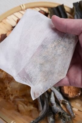 A hand holding up a homemade dashi packet over a bamboo tray with dashi ingredients