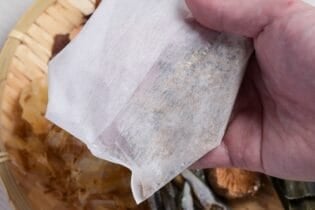A hand holding up a homemade dashi packet over a bamboo tray with dashi ingredients