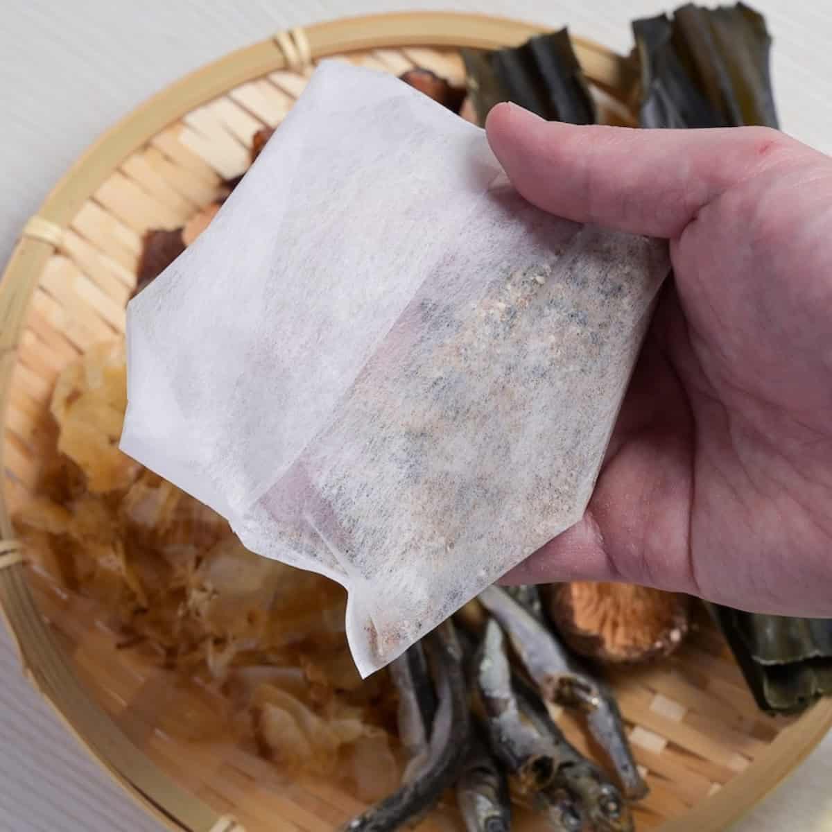 A hand holding up a homemade dashi packet over a bamboo tray with dashi ingredients