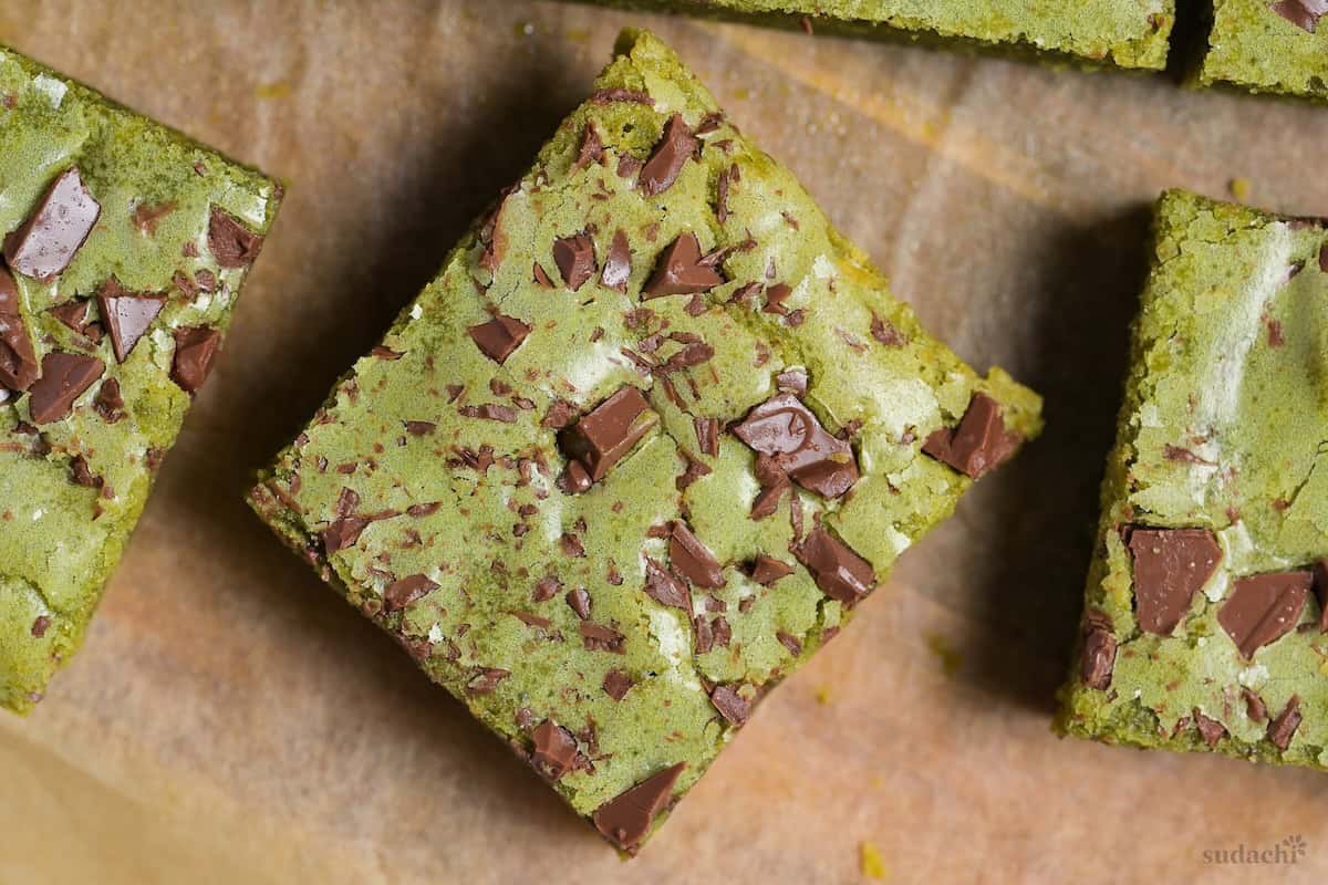 Close up of Matcha blondies topped with chunks of roughly chopped chocolate on a piece of brown parchment paper on a wooden cutting board