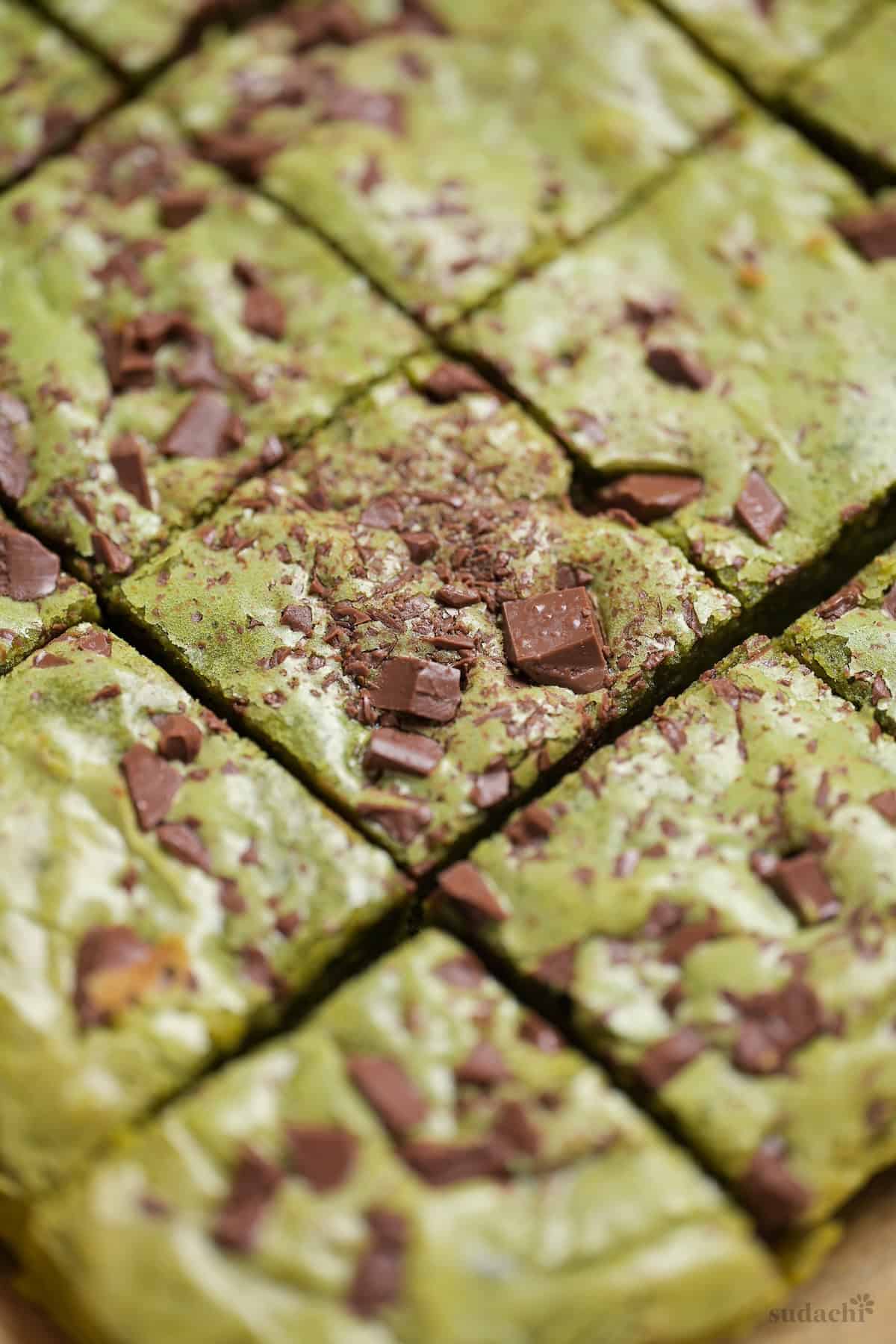 Matcha blondies topped with chunks of roughly chopped chocolate on a piece of brown parchment paper on a wooden cutting board