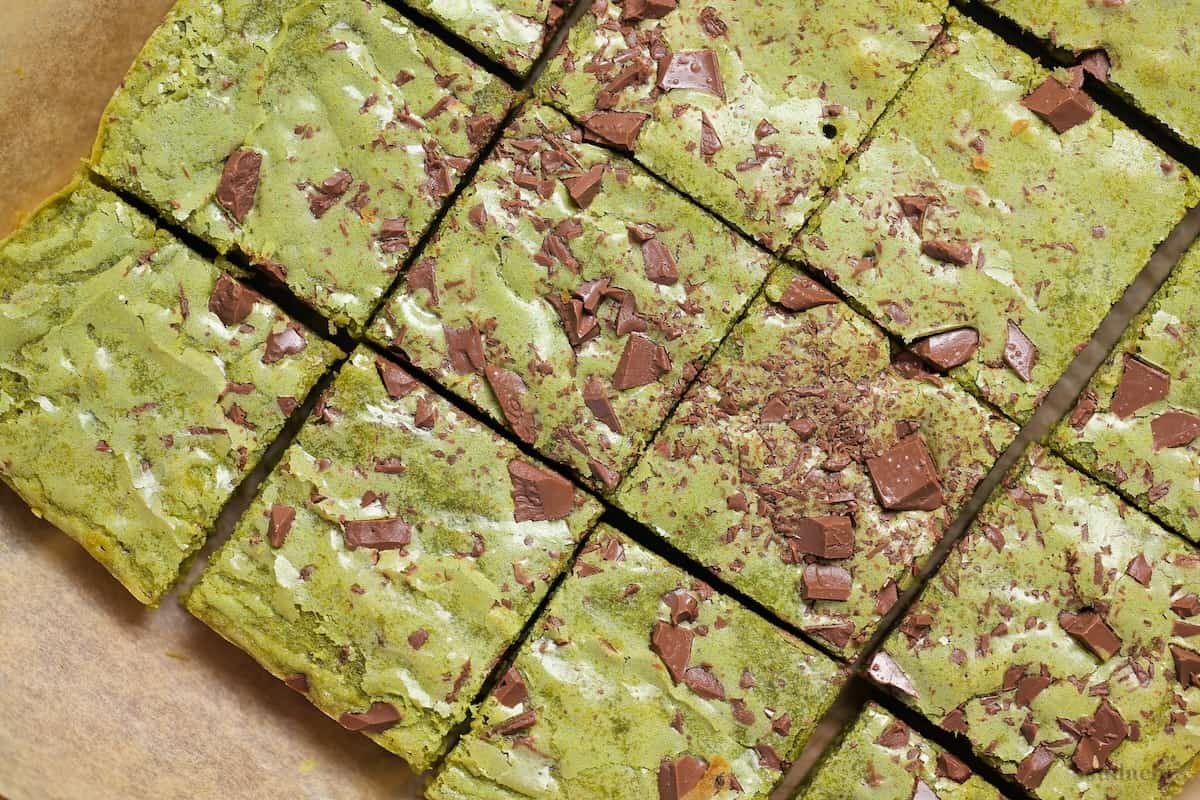 Matcha blondies topped with chunks of roughly chopped chocolate on a piece of brown parchment paper on a wooden cutting board