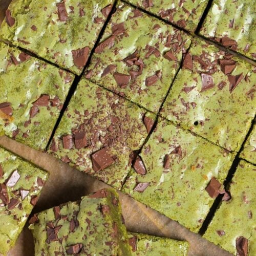 Matcha blondies topped with chunks of roughly chopped chocolate on a piece of brown parchment paper on a wooden cutting board