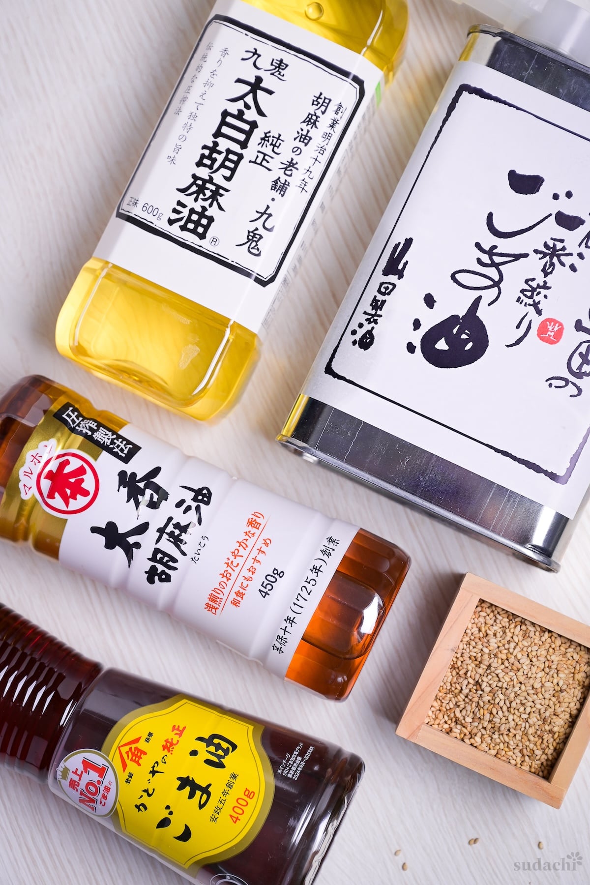 Various brands of bottled sesame oil arranged on a light wood-effect background next to a small masu filled with sesame seeds