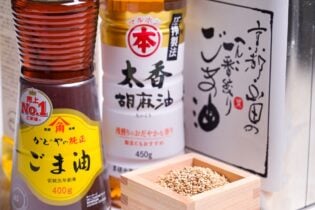 Various brands of bottled sesame oil arranged on a light wood-effect background next to a small masu filled with sesame seeds