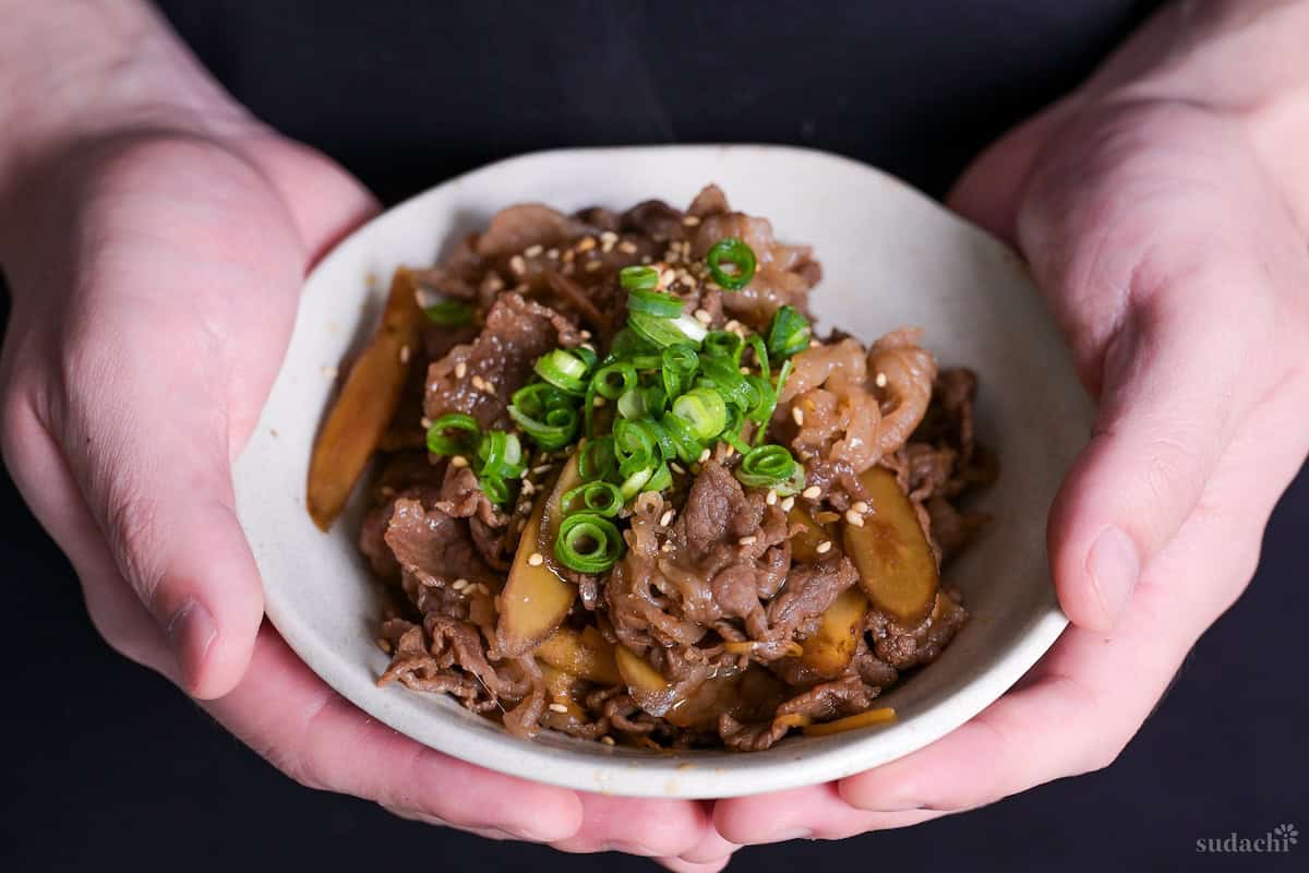 Yuto Omura holding up beef shigureni topped with chopped green onions and sesame seeds in a small off-white bowl