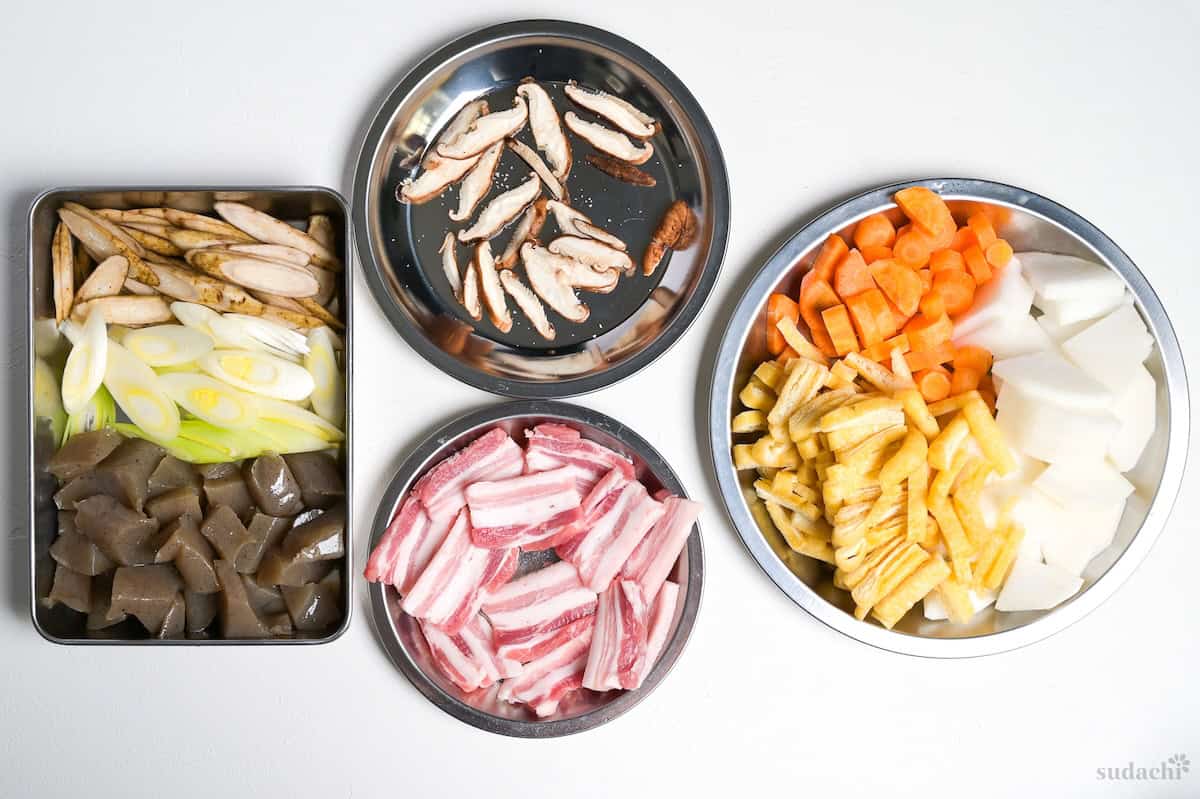 Prepared ingredients for making tonjiru. From left to right, thinly sliced burdock root, Japanese leek and roughly cut konnyaku, thinly sliced shiitake mushrooms, thick slices of pork belly, carrot half moons, daikon radish sliced and quartered, sliced tofu pouches (aburaage).