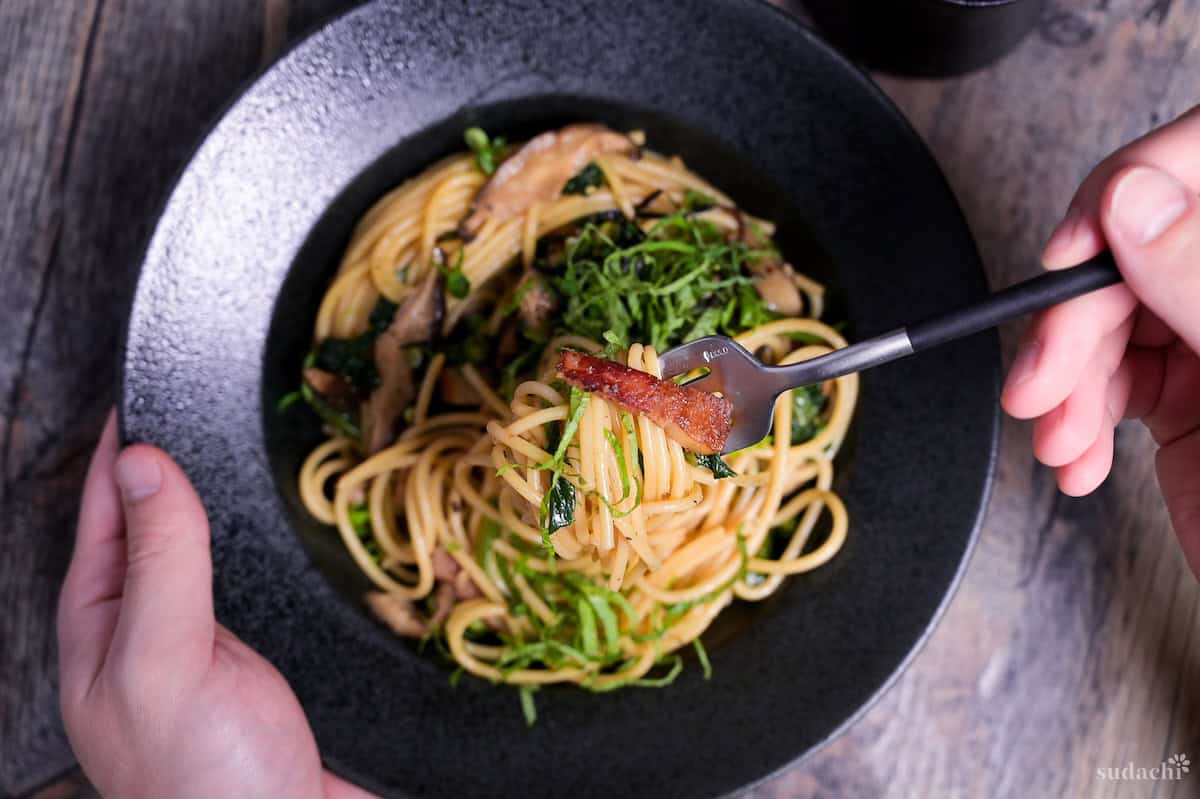 Japanese bacon and mushroom shoyu butter wafu pasta topped with shredded shiso leaves and nori served in a black pasta dish with a fork lifting some spaghetti and bacon.